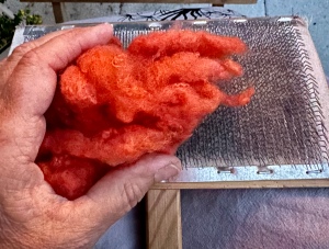 Eine Hand hält orange Wollfasern auf eine Drahtbürste.